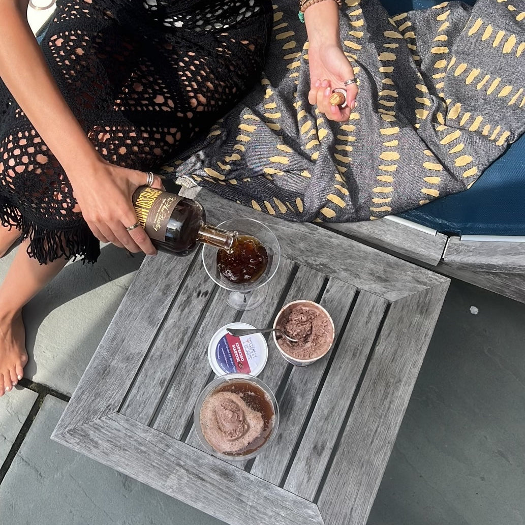 Person in a straw hat pouring an espresso martini into a glass  next to two pints of ice cream on a small table by a poolside.