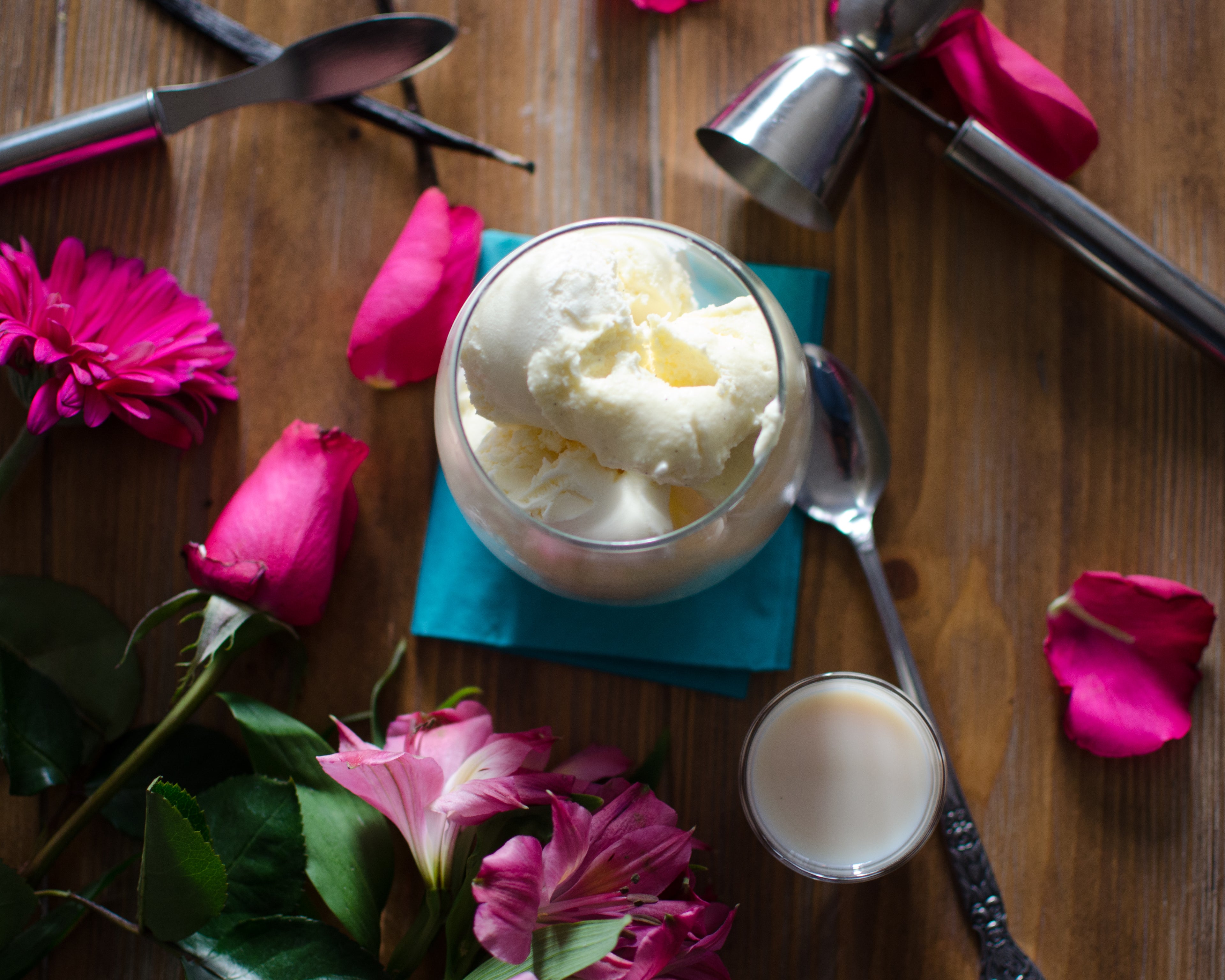 Vanilla ice cream in a glass bowl with flowers and spoons on a wooden table with pint roses and a shot glass