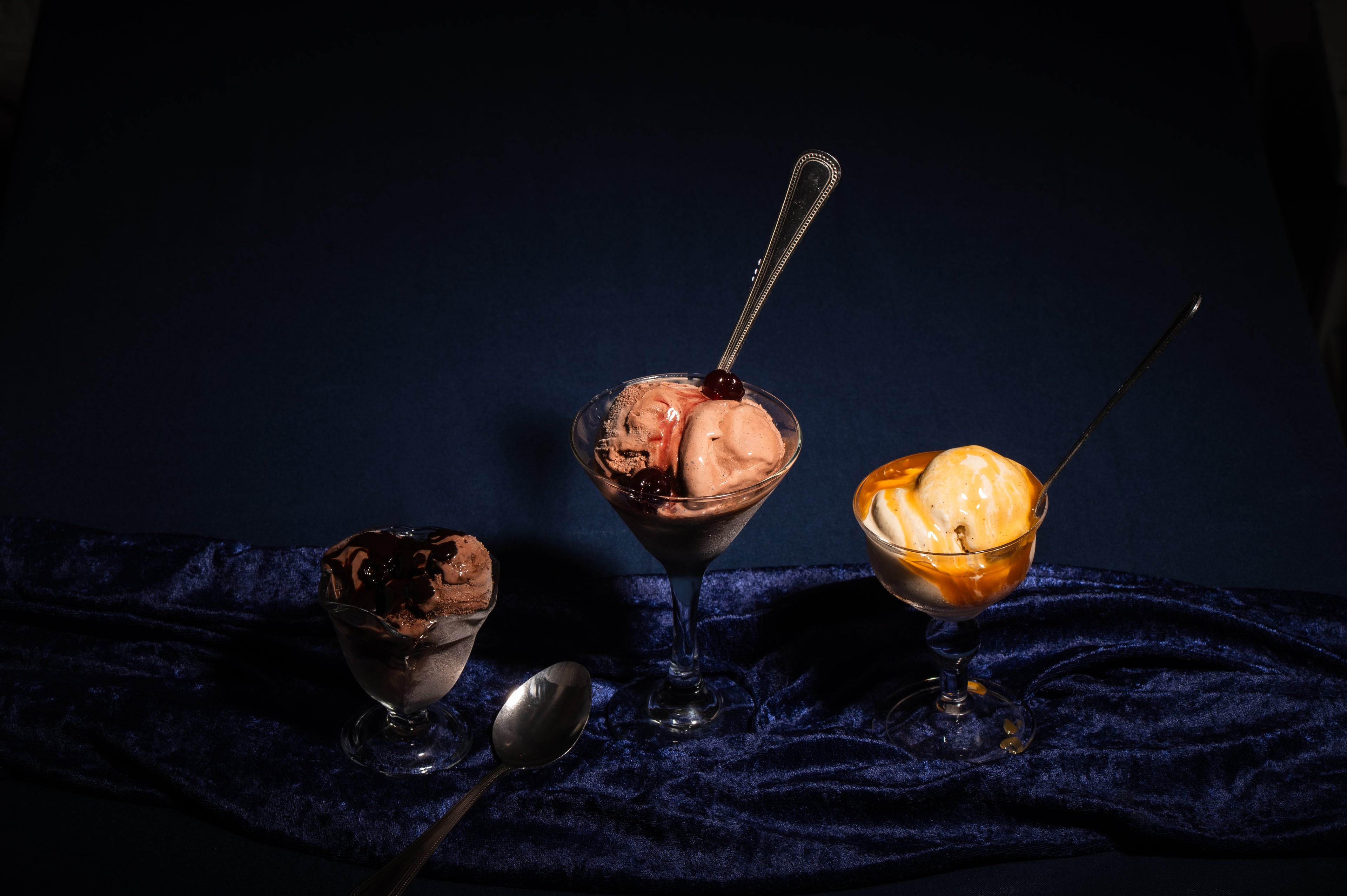 Three dessert glasses with boozy frozen custard and toppings on a dark blue textured surface