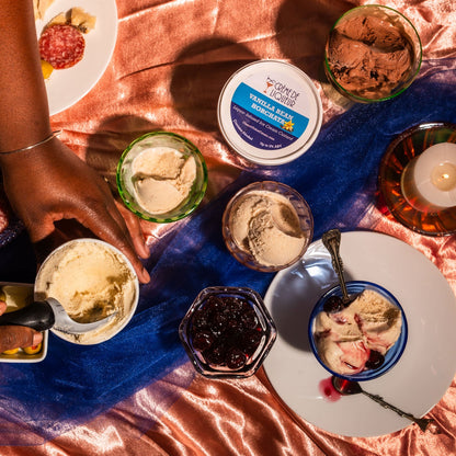People enjoying alcoholic ice cream at a table with various desserts and drinks on a pink blanket.